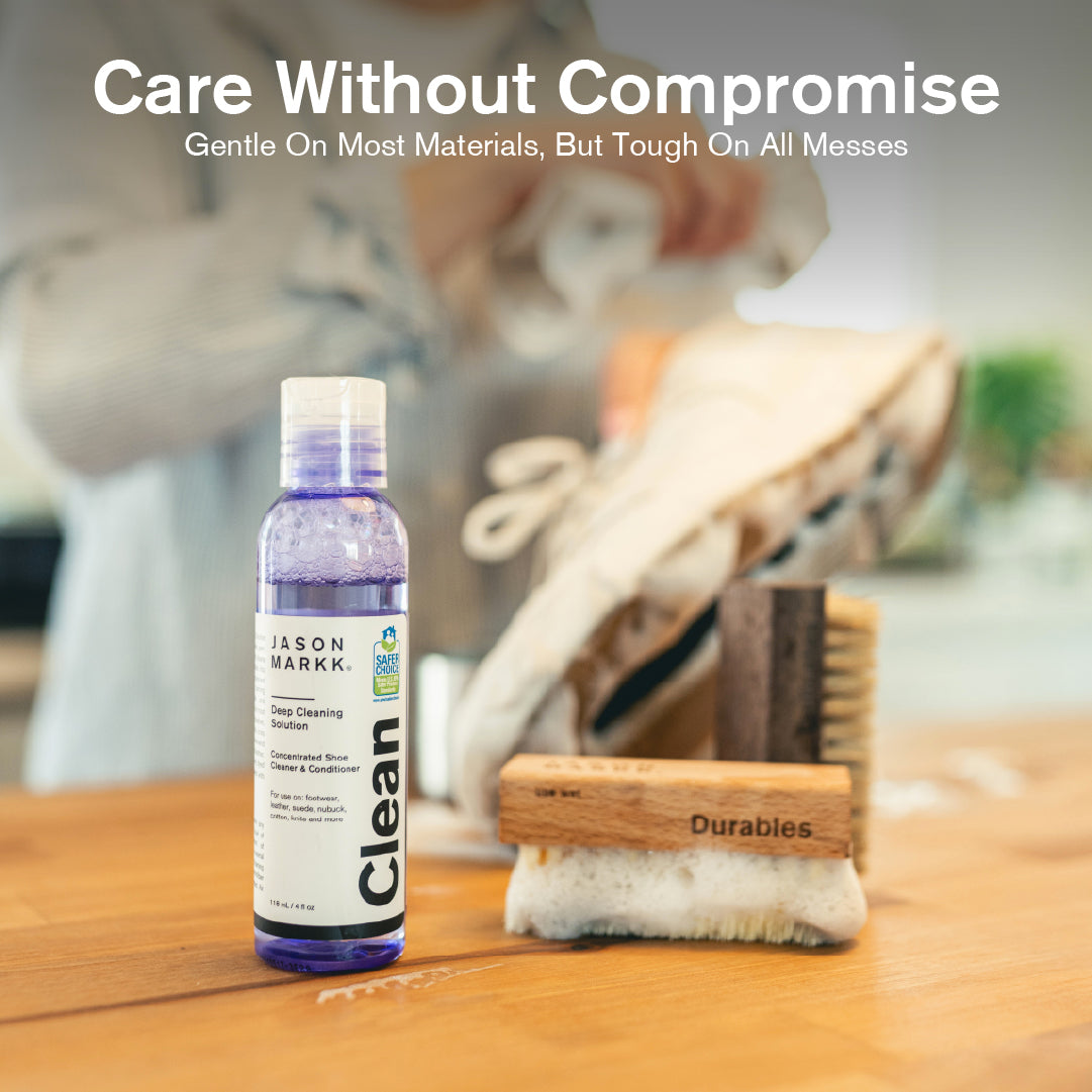 A 4 fl oz bottle of Jason Markk Deep Cleaning Solution concentrated shoe cleaner and conditioner in a purple bottle (EPA Safer Choice certified) displayed alongside two Jason Markk Durables wooden shoe cleaning brushes with soft and stiff bristles on a wood table. A person cleaning a white canvas sneaker is visible in the blurred background. Text overlay reads: "Care Without Compromise – Gentle On Most Materials, But Tough On All Messes."