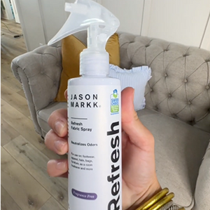 Hand holding a bottle of Jason Markk Refresh n a living room setting.